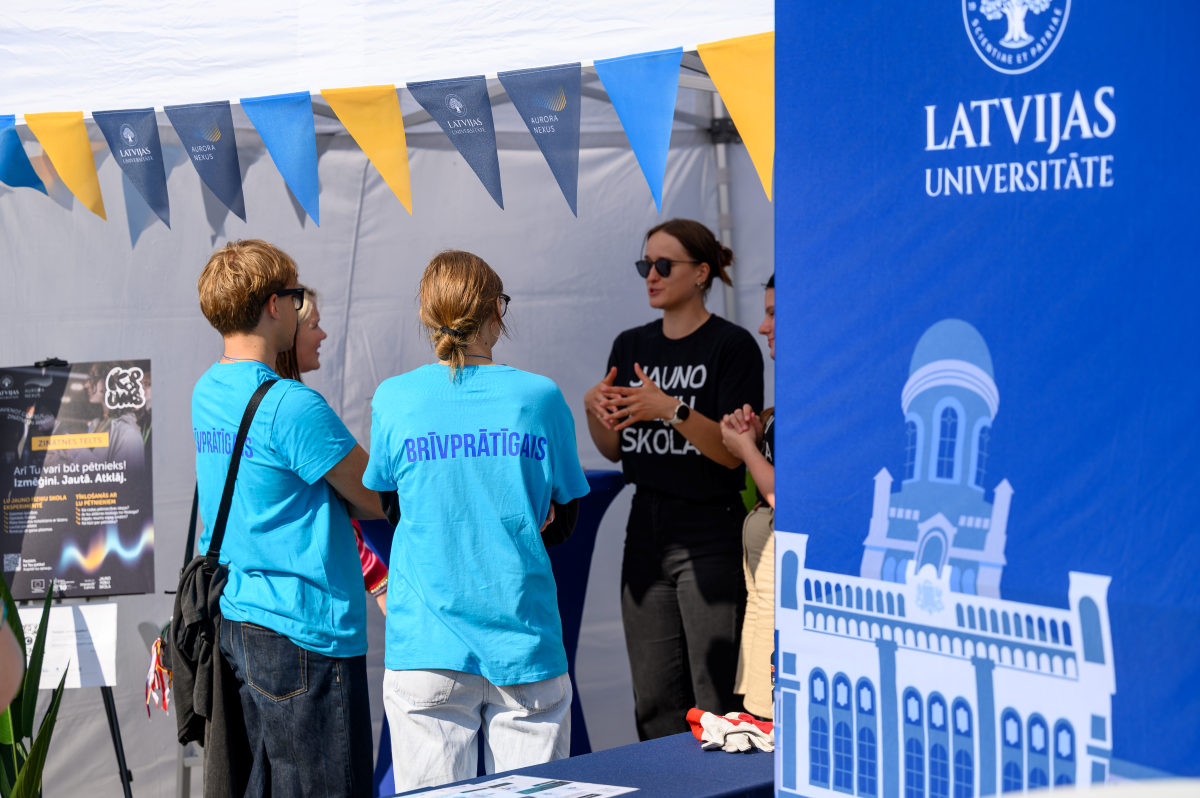 Jauniešu iespēju festivāls "KOPUMS 2026". Foto: Ritvars Pujats Brīvprātīgie un pasākuma dalībnieki sarunājas Latvijas Universitātes teltī. Priekšplānā redzams Latvijas Universitātes stends, bet virs telts izvietoti LU un Aurora Nexus karodziņi.