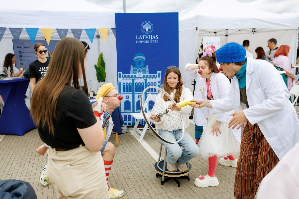 Jauniešu iespēju festivāls "KOPUMS 2026". Foto: Toms Norde, LU Bērni un pieaugušie piedalās zinātnes aktivitātē Latvijas Universitātes teltī. Dalībnieks griež velosipēda riteni, kamēr laboratorijas halātos tērpti pasākuma vadītāji demonstrē eksperimentu. Fonā redzams Latvijas Universitātes stends.