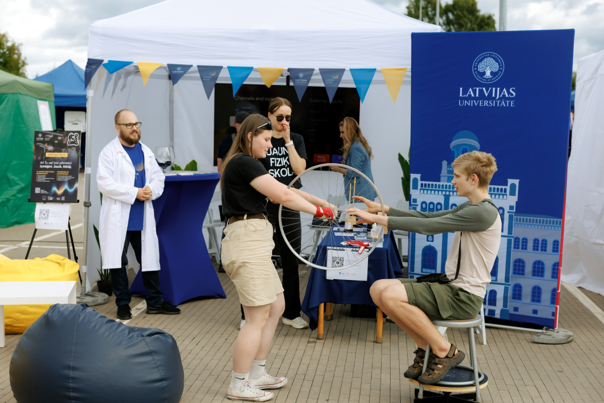 Jauniešu iespēju festivāls "KOPUMS 2026". Foto: Toms Norde, LU Jaunieši piedalās praktiskā zinātnes aktivitātē pie Latvijas Universitātes telts, griežot velosipēda riteni. Apkārt redzami pasākuma dalībnieki, LU stends un zinātnes demonstrējumu aprīkojums.