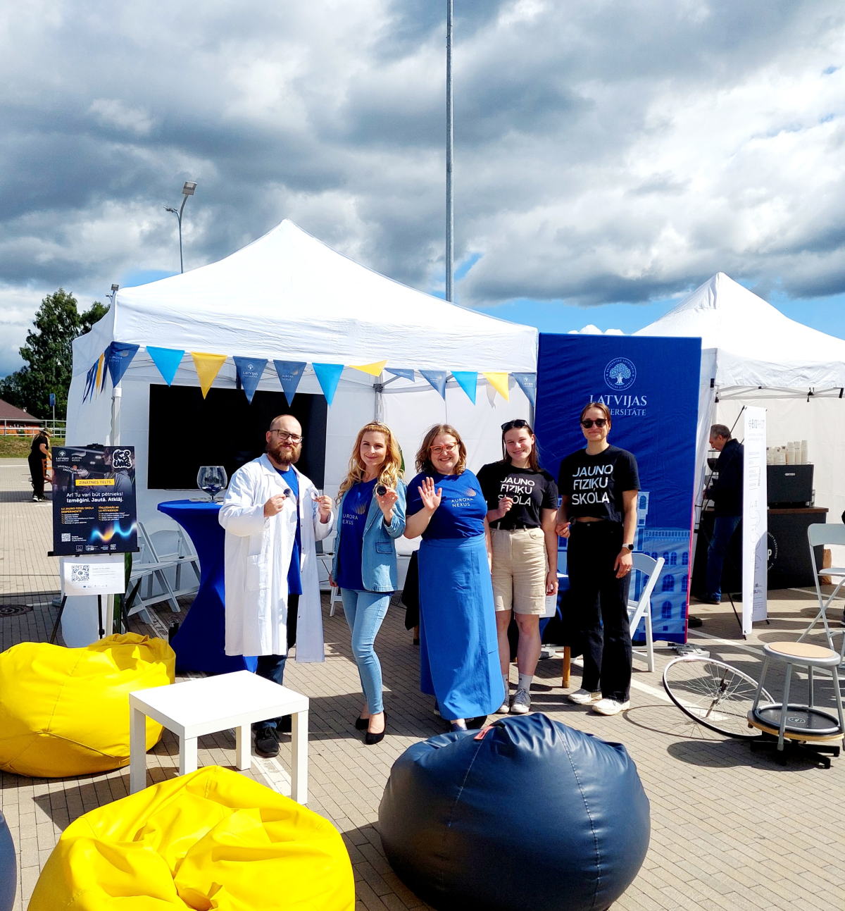 Jauniešu iespēju festivāls "KOPUMS 2026". Foto: "Aurora Nexus" foto arhīvs Latvijas Universitātes zinātnes telts komanda pozē kopbildei pasākumā brīvā dabā. Priekšplānā redzami sēžammaisi, bet fonā Latvijas Universitātes stends un aktivitāšu telts.