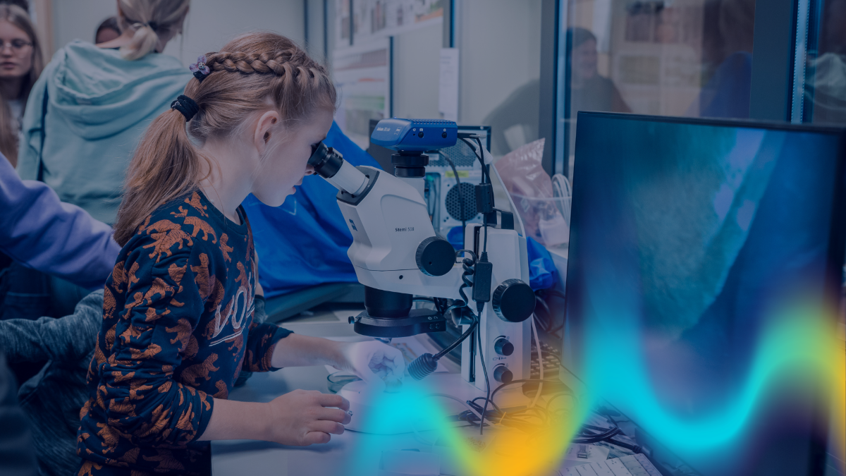 Apmeklētājs sēž pie laboratorijas galda un raugās mikroskopā. Blakus novietots dators ar palielinātu parauga attēlu ekrānā, bet fonā redzami citi pasākuma dalībnieki zinātnes demonstrāciju telpā.